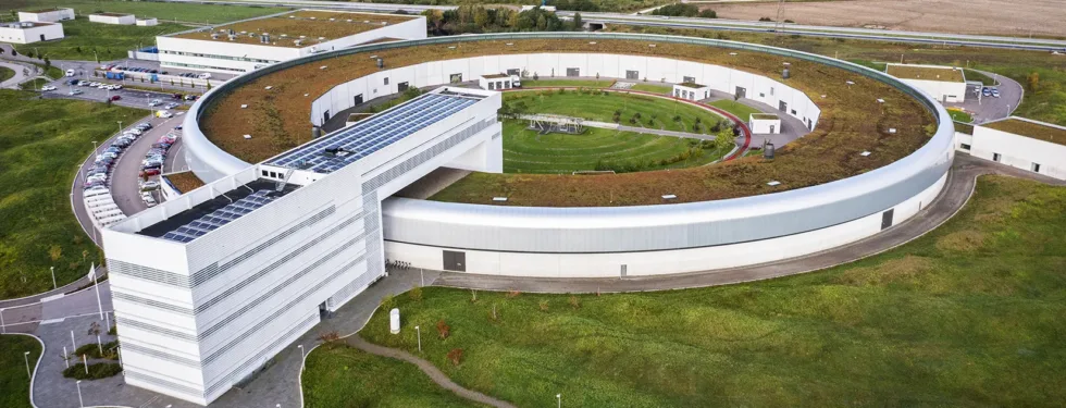 Drone image of MAX IV from above, surrounded by green landscape and the horizon.